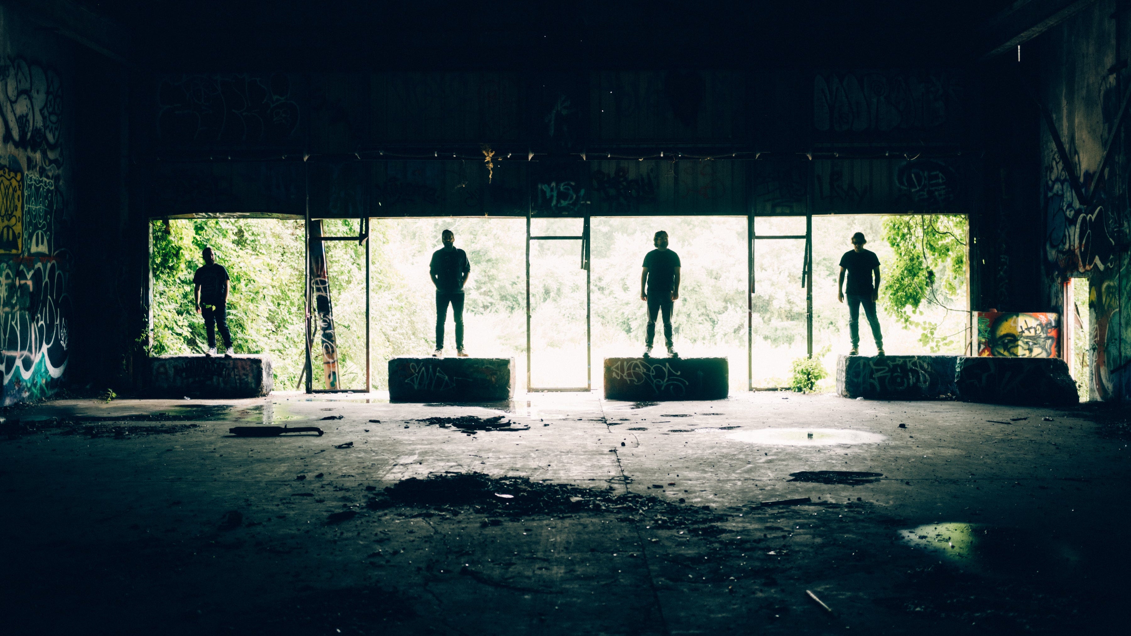 Four silhouettes standing in front of large windows in an abandoned building.